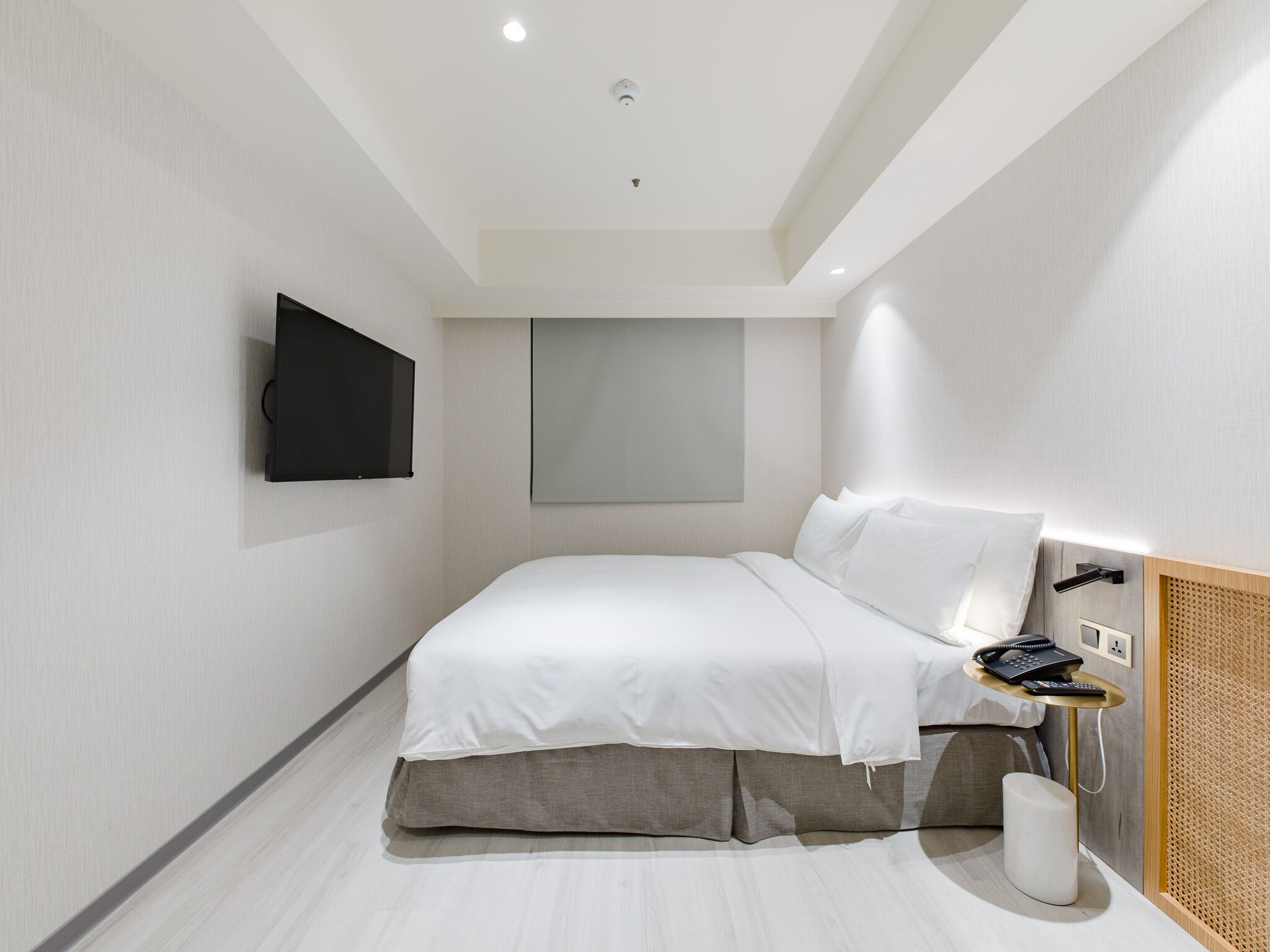 A modern bedroom with a bed, a mounted TV, a bedside table with a phone, and a small stool.