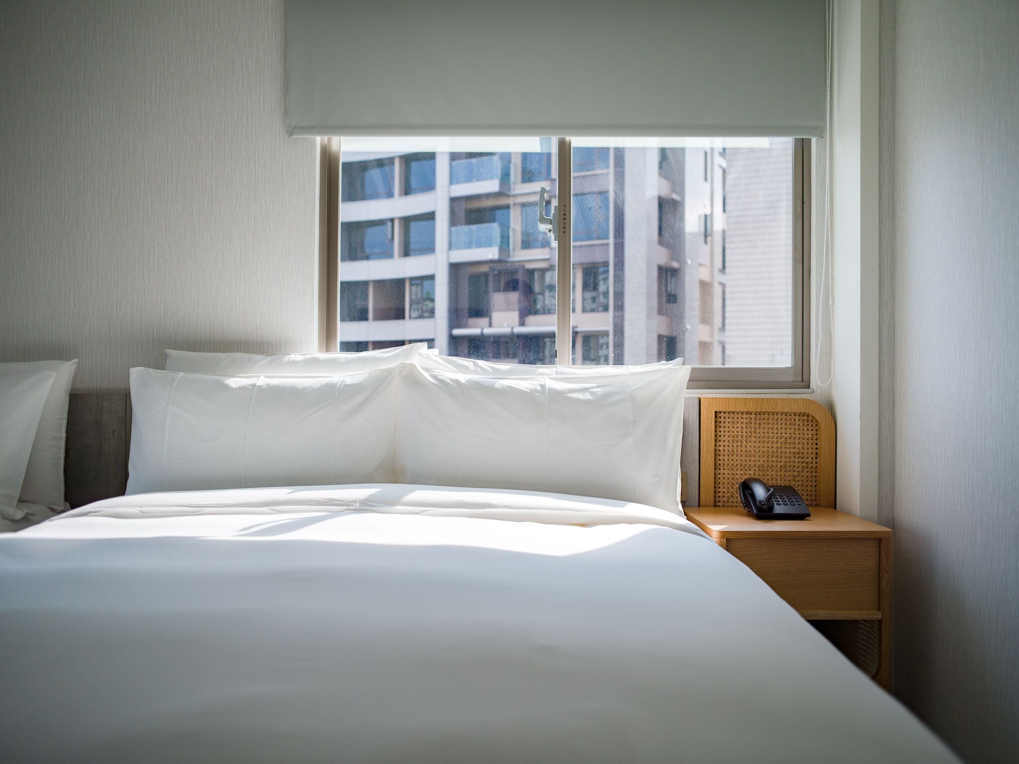 Habitación de hotel con una cama grande, una mesita de noche con teléfono y ventana con vista a edificios.