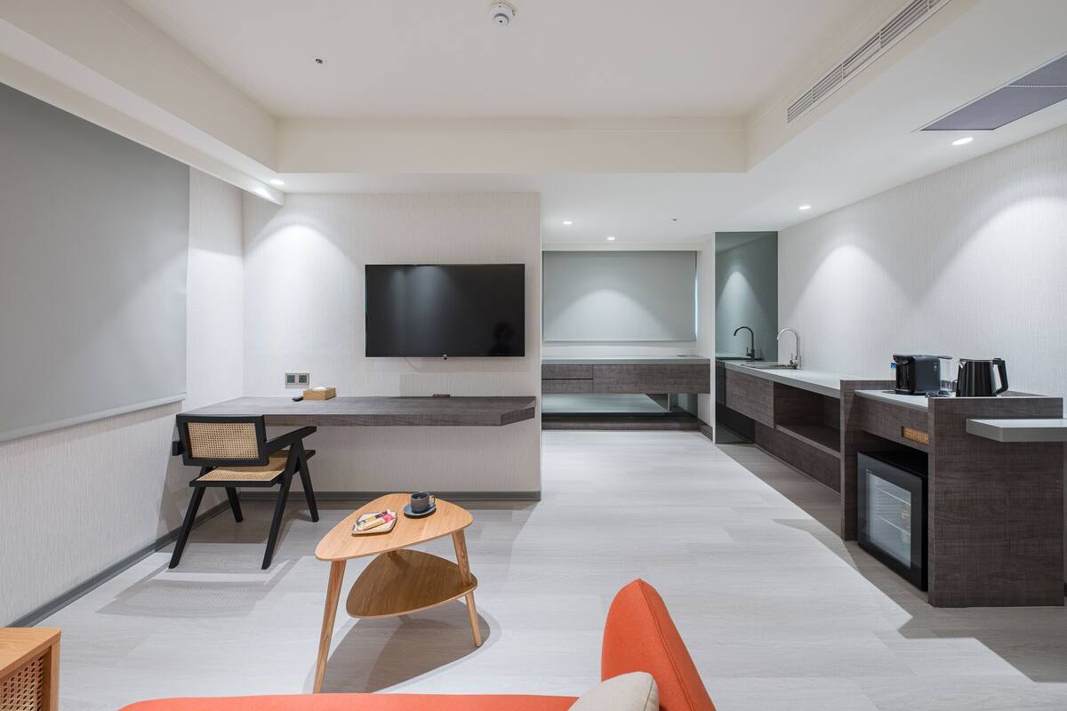 A modern hotel room with a flat-screen TV, a dining area with a table and chairs, a kitchenette, and a sofa.