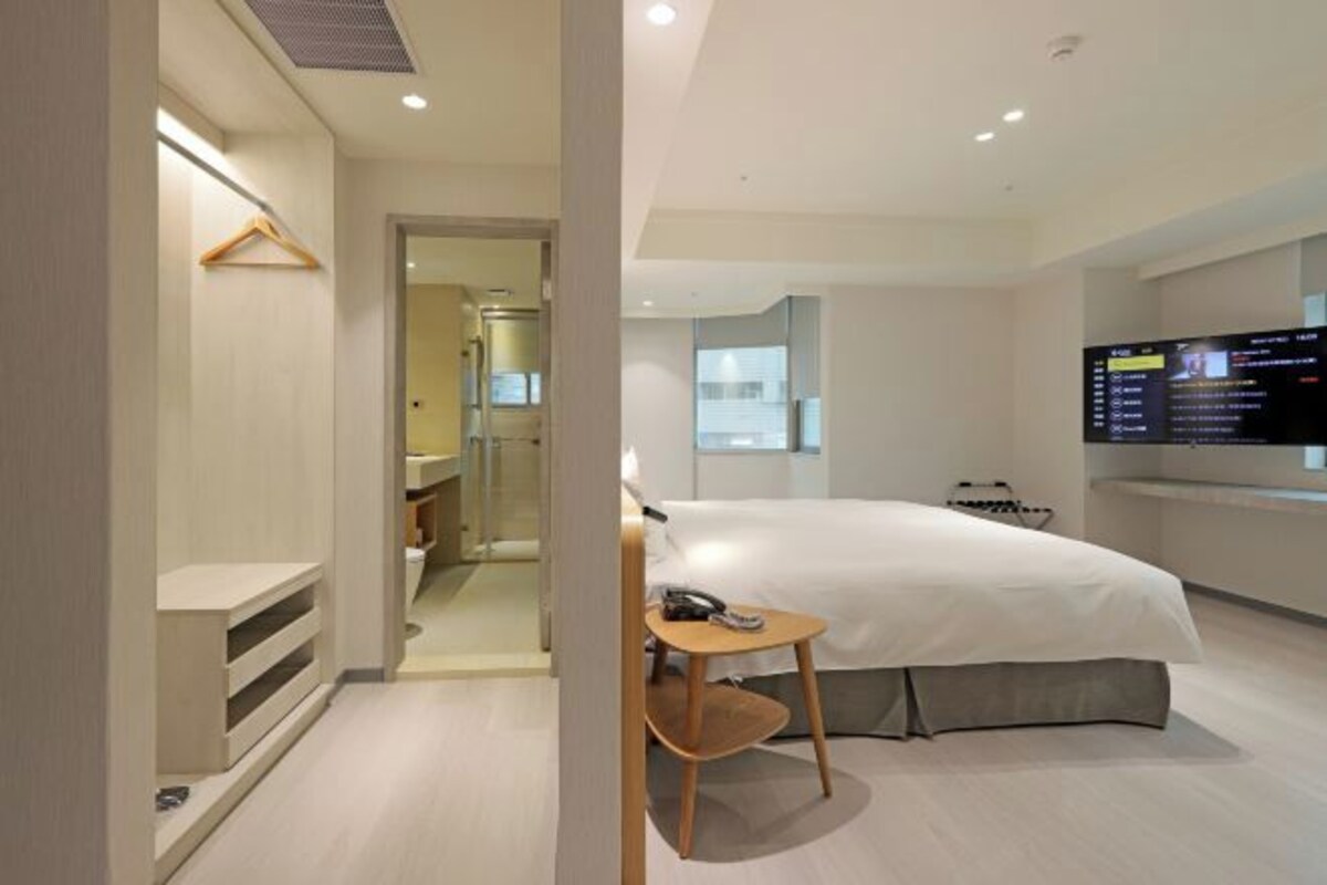 Una habitación de hotel moderna con una cama grande, una mesita de noche, un televisor montado en la pared y un vestidor.