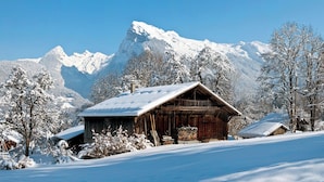 Eine Holzhütte mit schneebedecktem Dach, umgeben von schneebedeckten Bäumen und Bergen im Hintergrund.