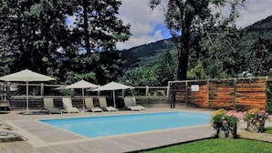 Ein rechteckiger Swimmingpool mit Liegestühlen und Sonnenschirmen, umgeben von einer Holzterrasse und einer Rasenfläche.