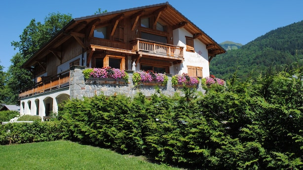 Ein Chalet mit Steinfassade und Holzbau, mit einem Balkon mit Blumenkästen und Blick auf eine Berglandschaft.