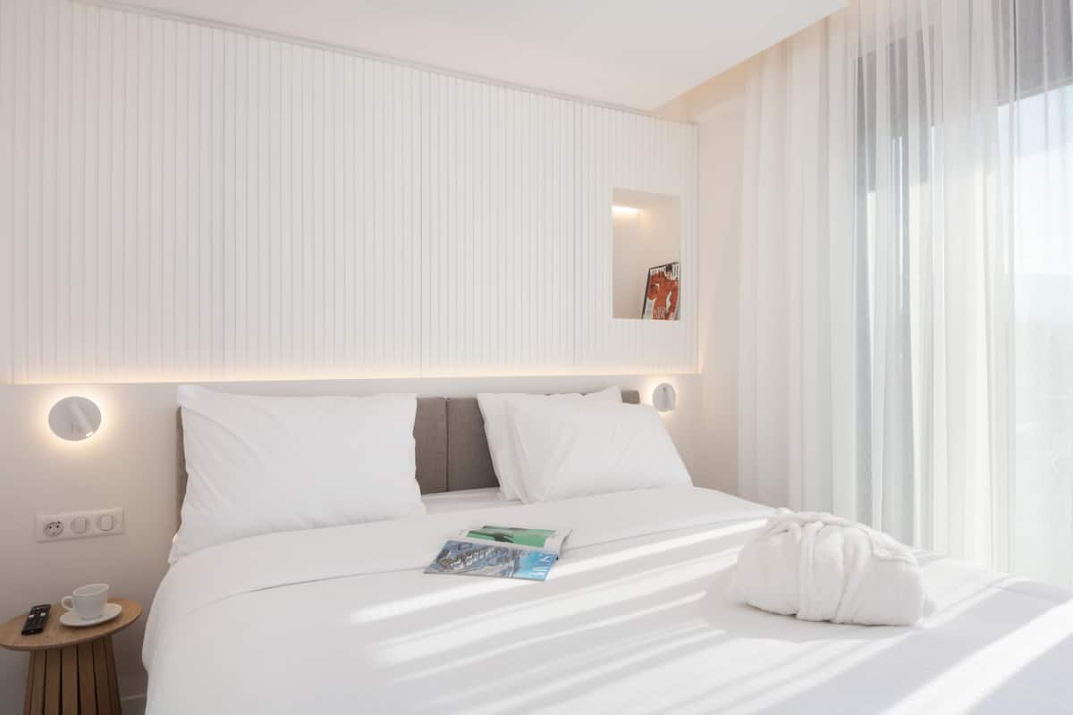 Habitación de hotel con una cama bien hecha, una mesita de noche con una taza y un control remoto, y una ventana con cortinas translúcidas.