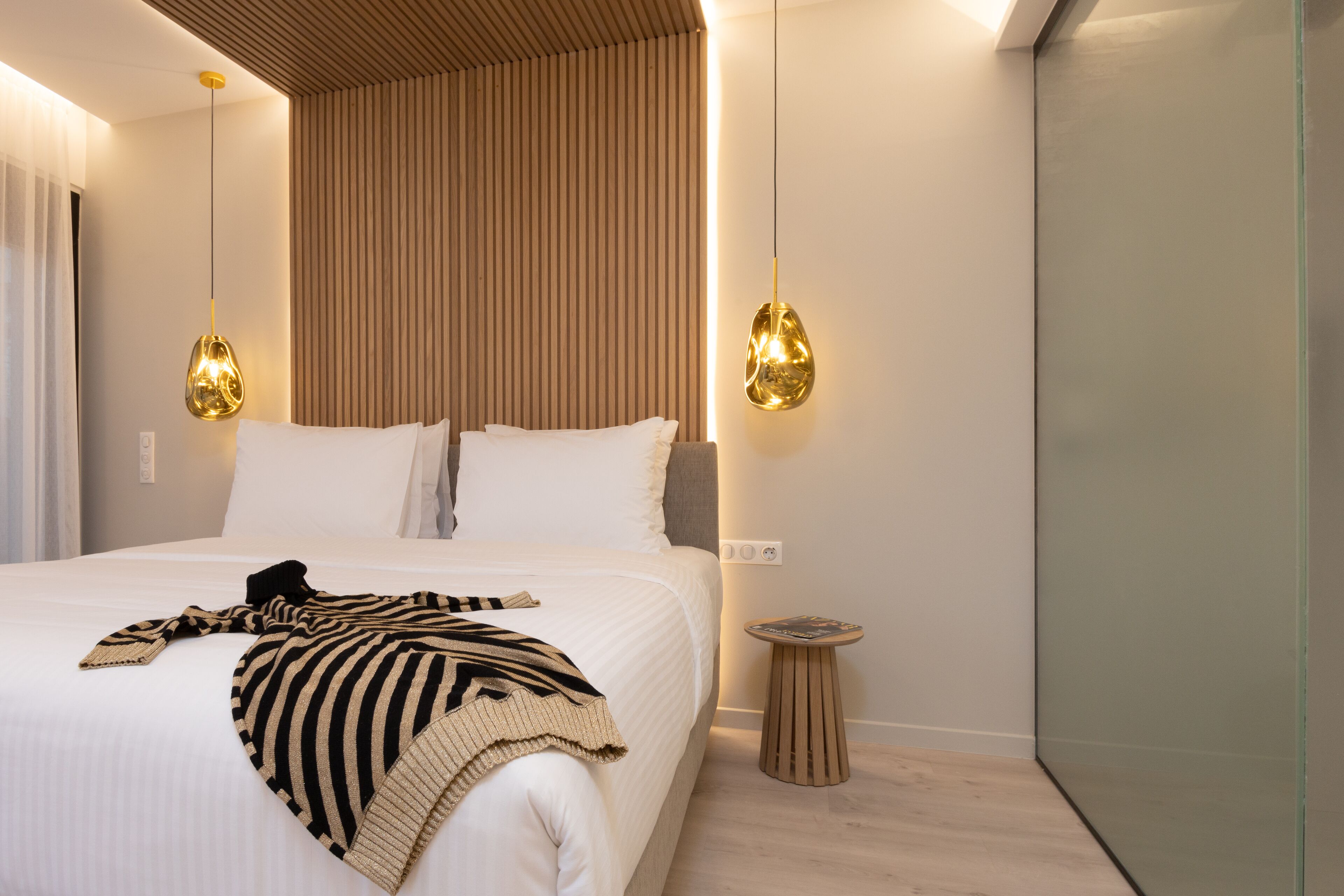 A modern hotel room with a large bed, two pendant lights, a small wooden side table, and a wall with vertical wooden slats.
