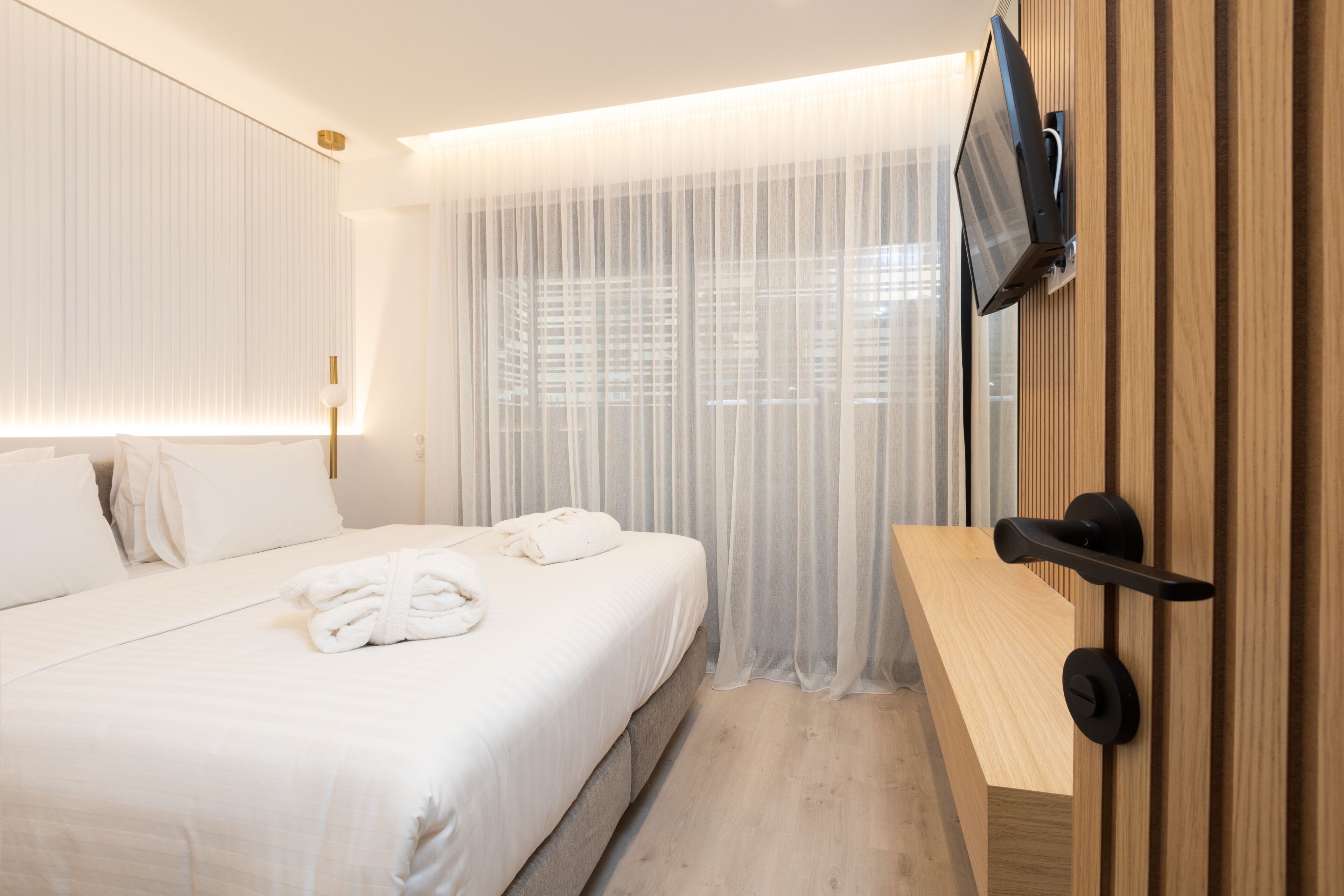 A hotel room with a large bed, a TV mounted on the wall, and sheer curtains.