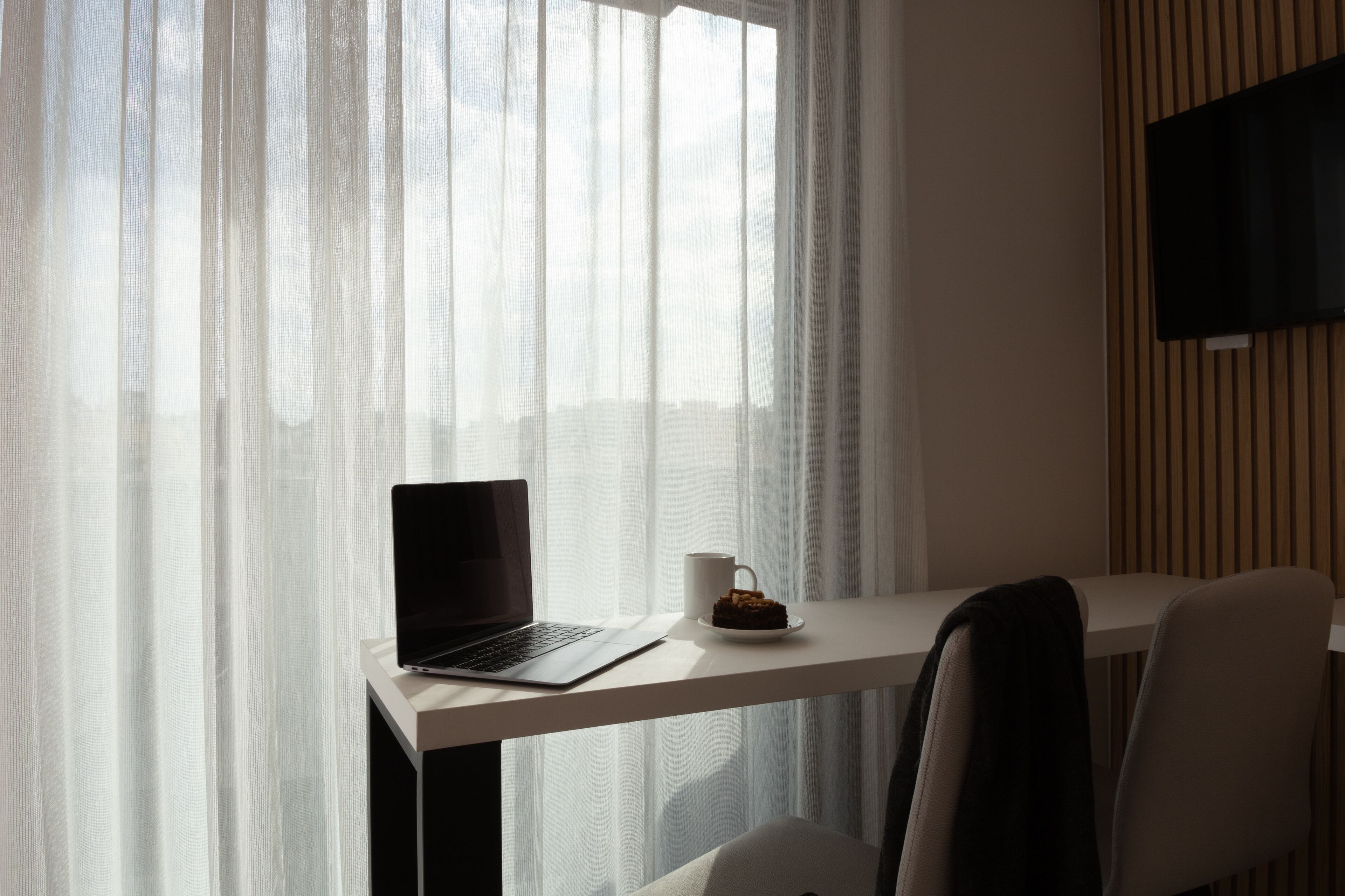 A room with a laptop on a desk, a cup of coffee, and a TV mounted on the wall.
