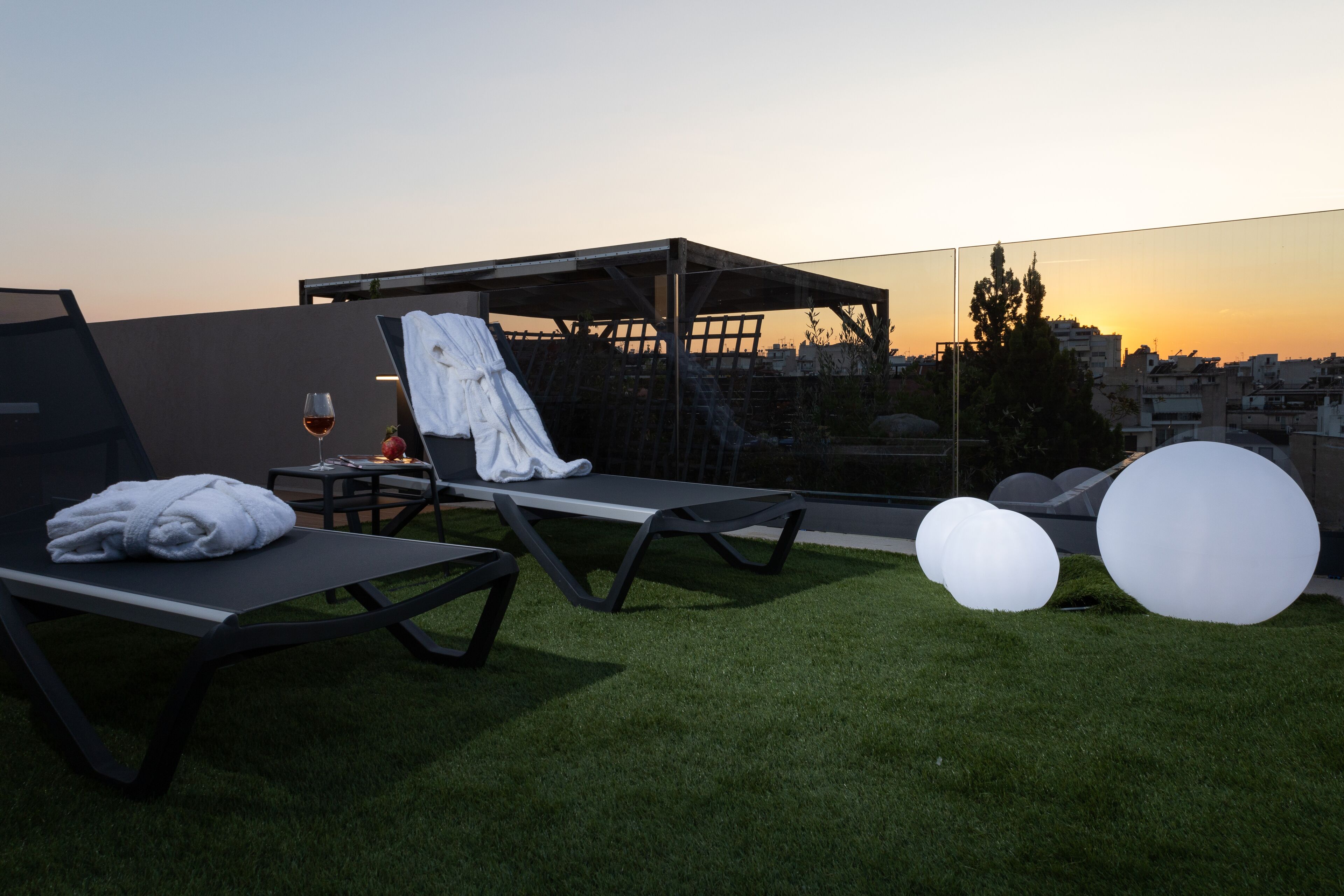 A rooftop lounge with sun loungers, a glass balustrade, and a sunset view.
