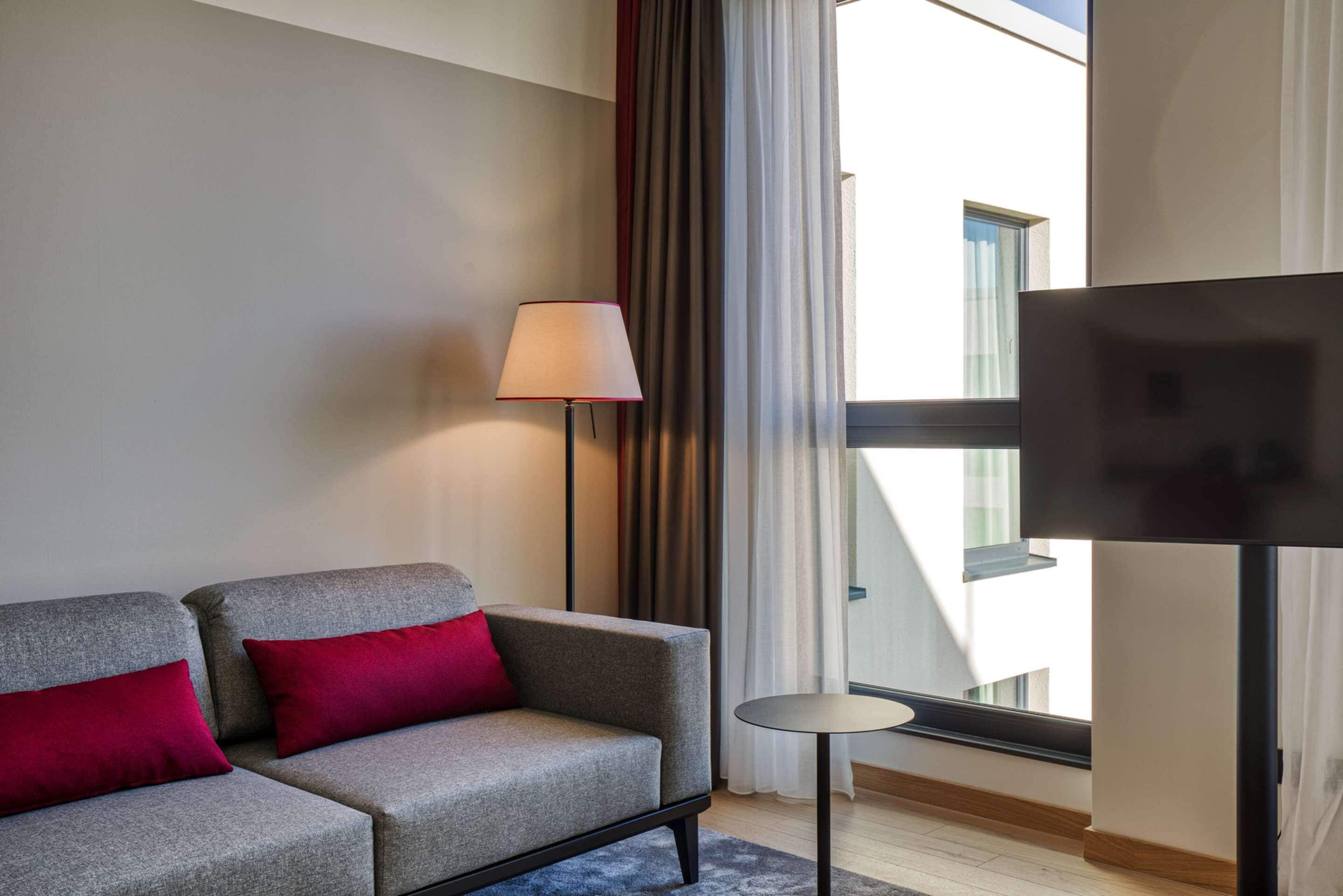 Una habitación de hotel moderna con un sofá gris, cojines rojos, una lámpara de pie, una pequeña mesa redonda y un televisor sobre un mueble bajo.