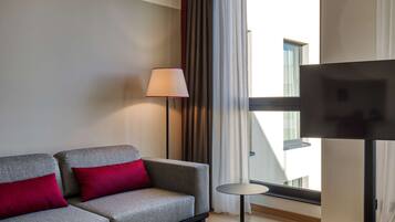 Una habitación de hotel moderna con un sofá gris, cojines rojos, una lámpara de pie, una pequeña mesa redonda y un televisor sobre un mueble bajo.