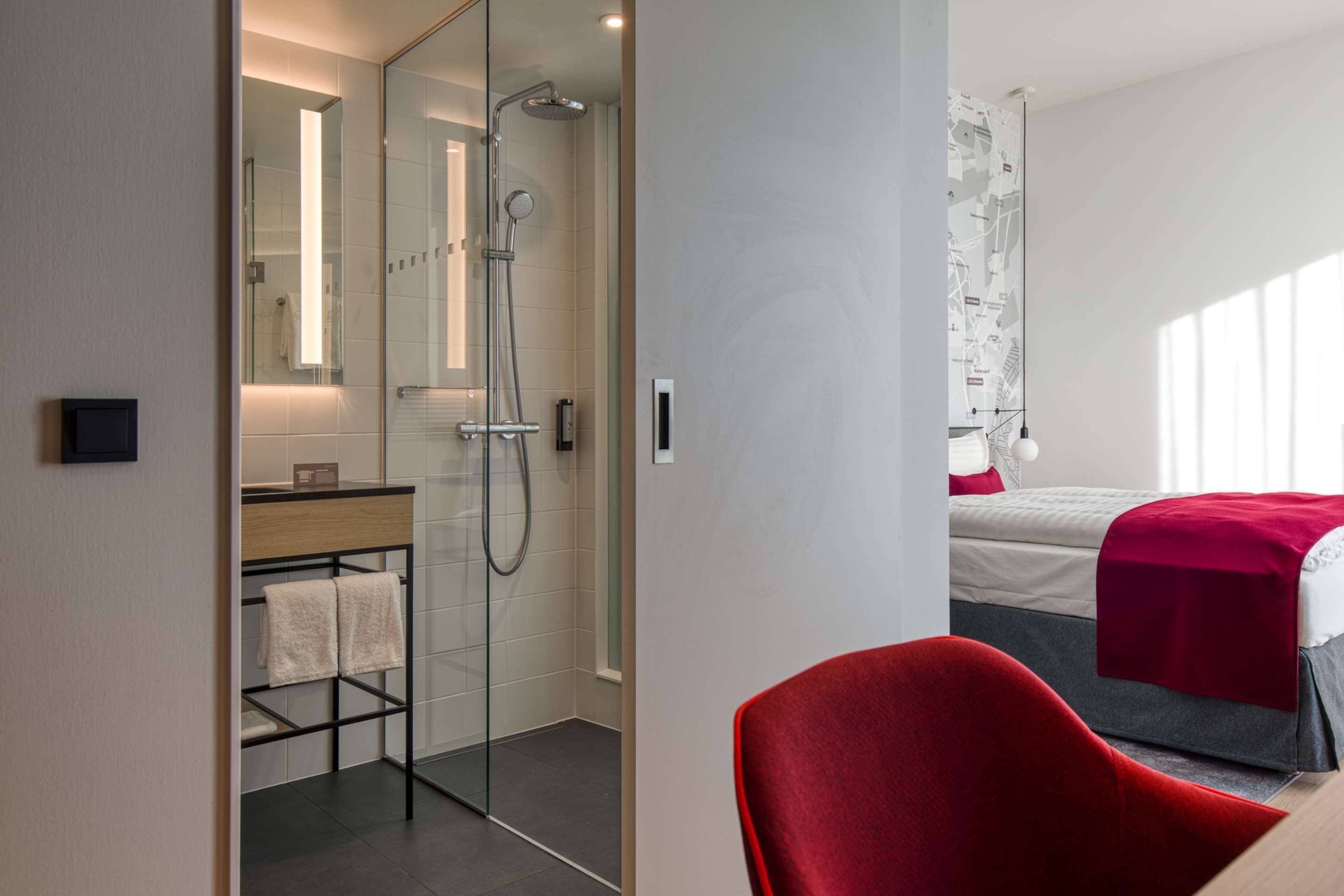 Een moderne hotelkamer met een glazen douchecabine, een rode stoel en een bed met een rode deken.