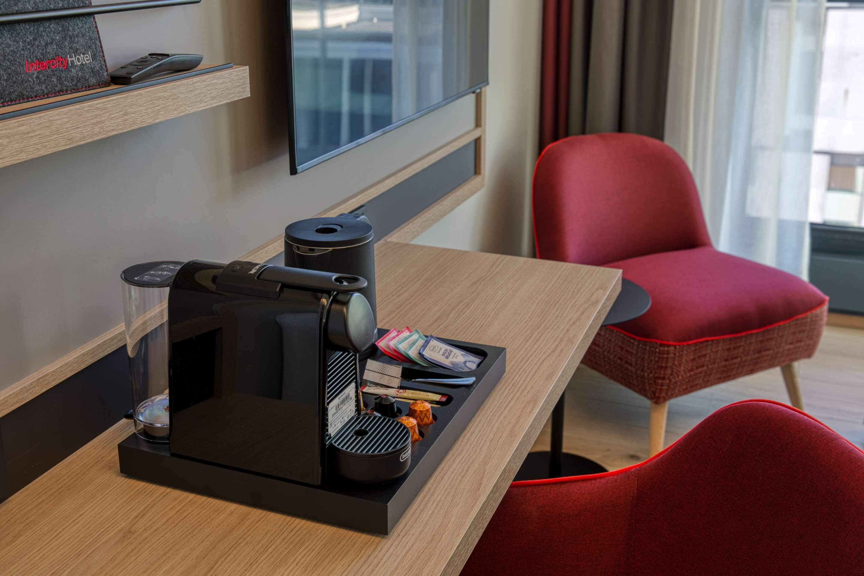 A compact hotel room with a modern coffee maker on the table, a flat-screen TV on the wall, and a red chair.