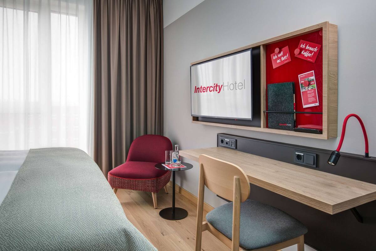 Habitación de hotel con cama, escritorio con silla, televisión y un tablón rojo con información del hotel.