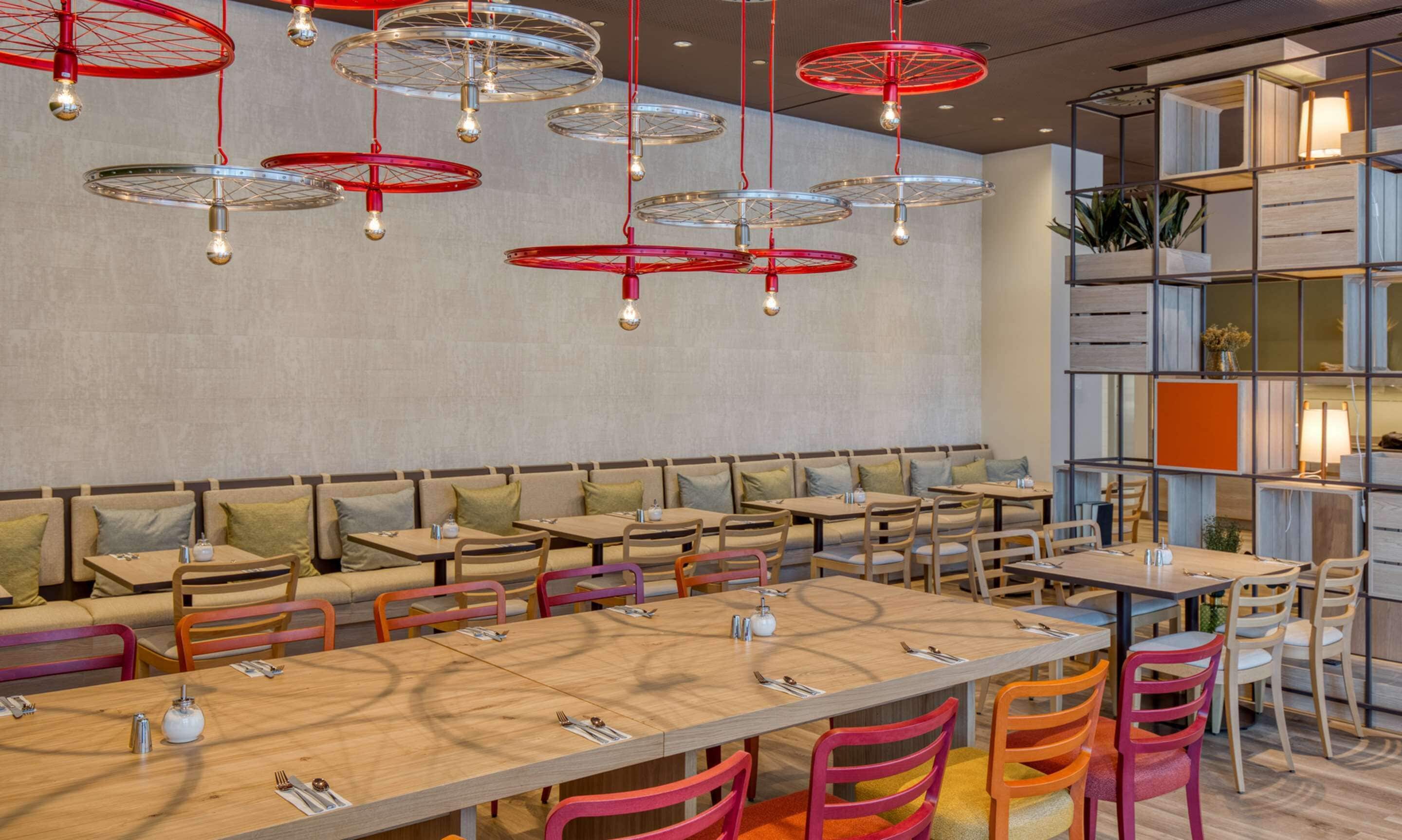 Un restaurante moderno con varias mesas y sillas, una zona de estar con un largo banco y una pared con una cuadrícula de estantes que contienen plantas y objetos decorativos.