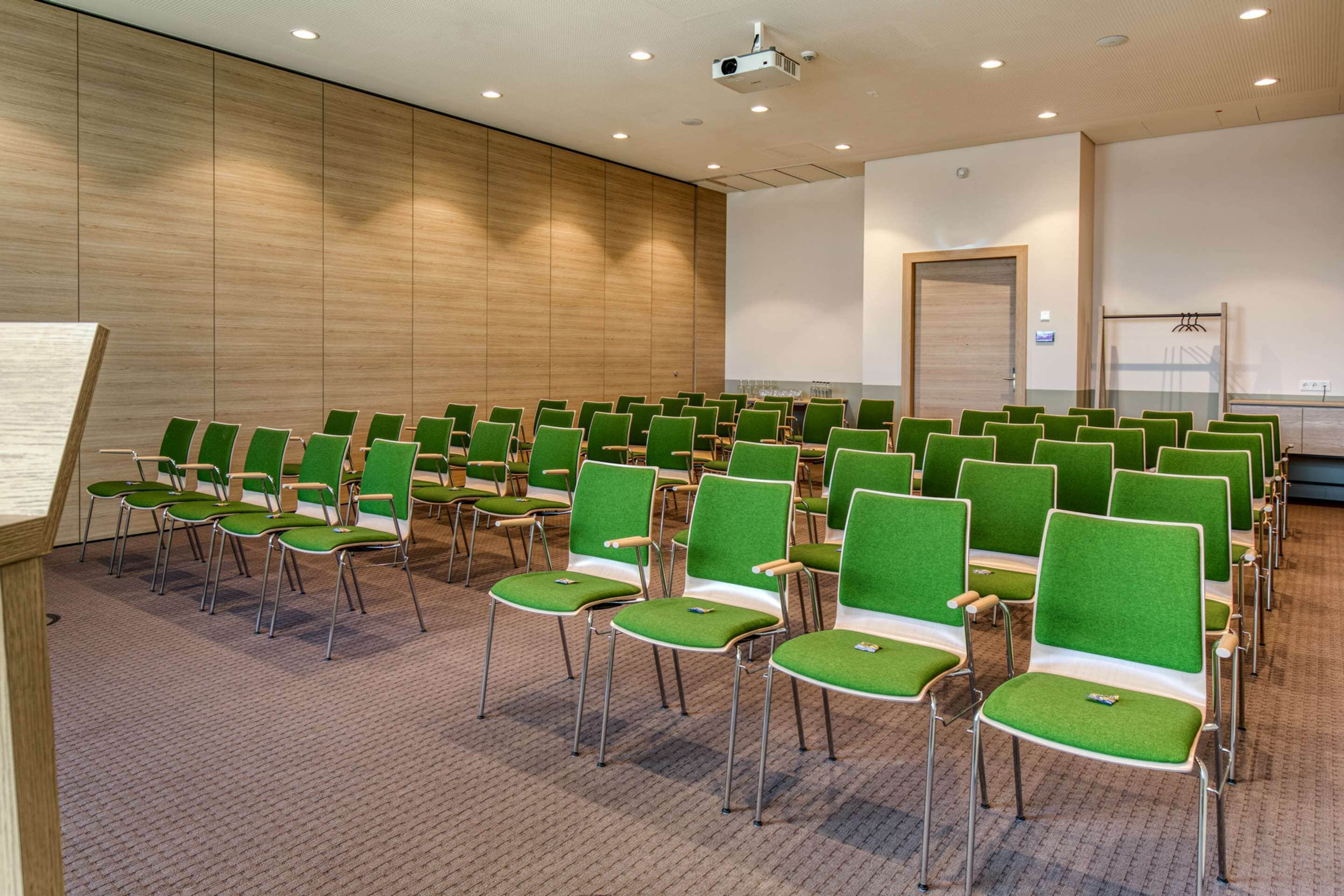 Une salle de conférence avec des rangées de chaises vertes, un écran de projection et un tableau blanc.
