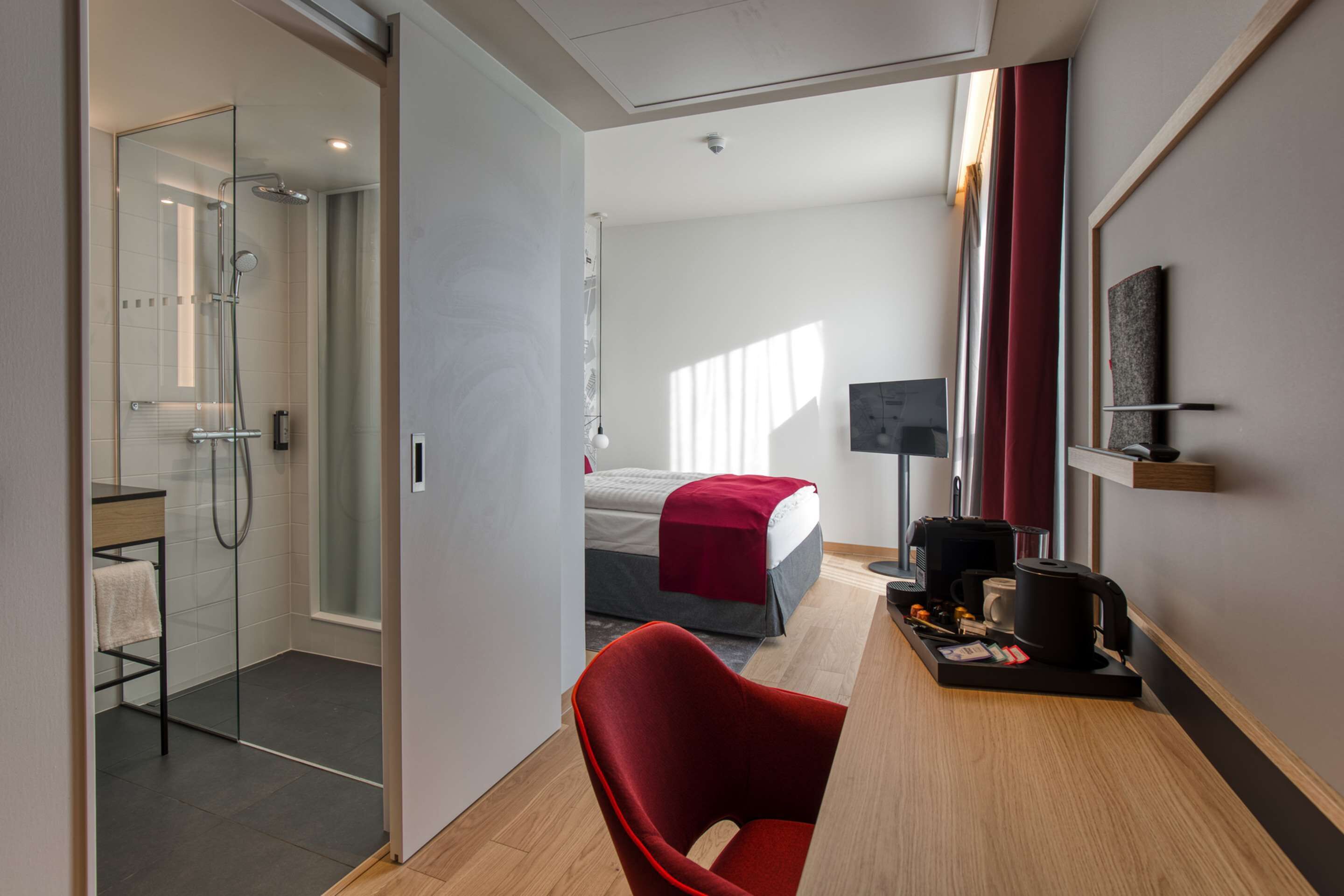 A modern hotel room with a glass shower enclosure, a bed with a red blanket, a red chair, a coffee machine, and a flat-screen TV on the wall.