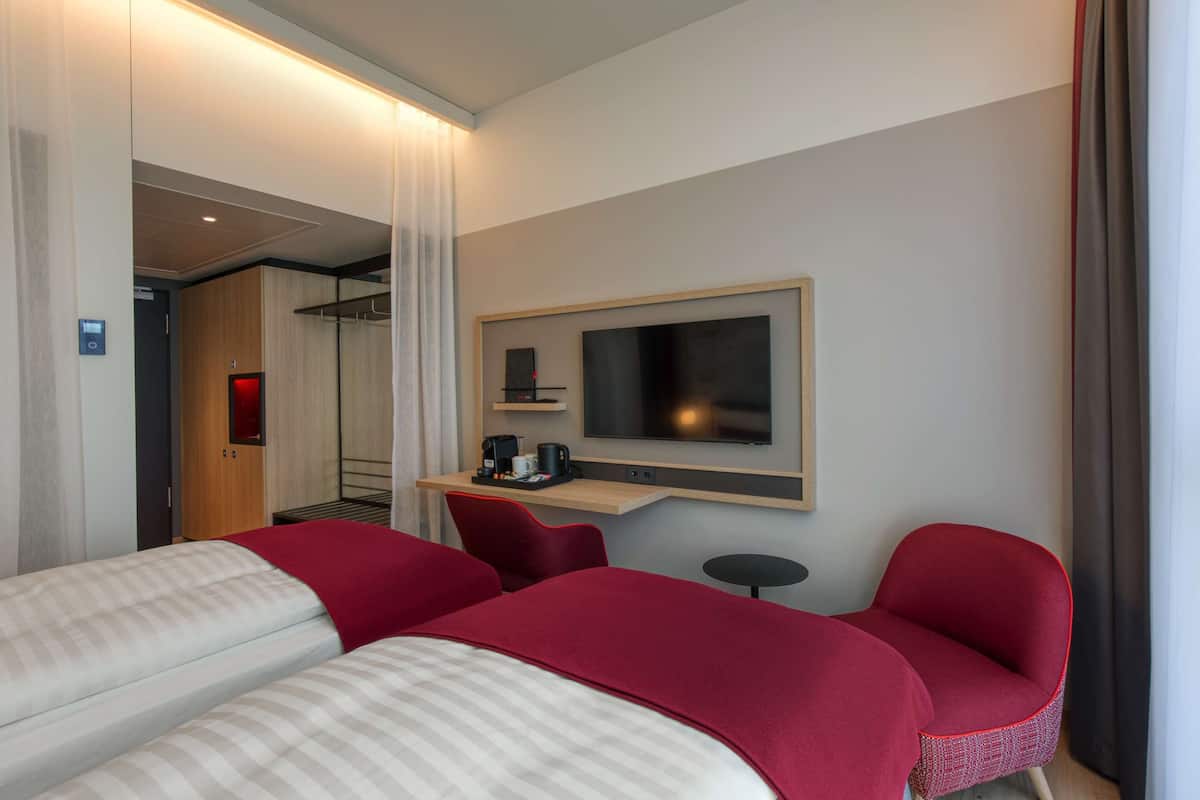 Una habitación de hotel con dos camas, televisión, una mesita con cafetera y una silla roja.