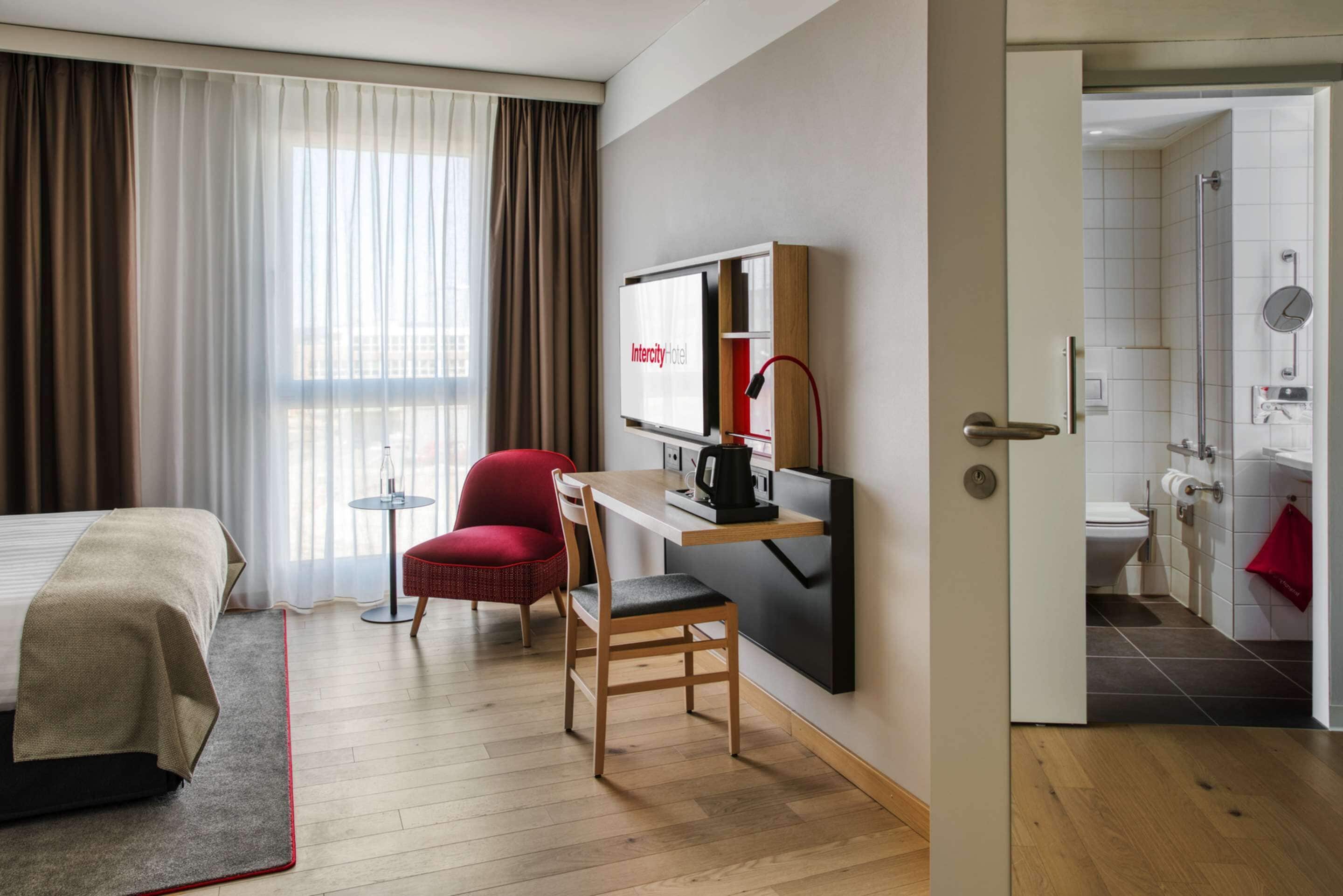 Une chambre d’hôtel équipée d’un lit, d’un bureau avec un ordinateur, d’une chaise et offrant une vue sur la ville grâce à de grandes fenêtres.