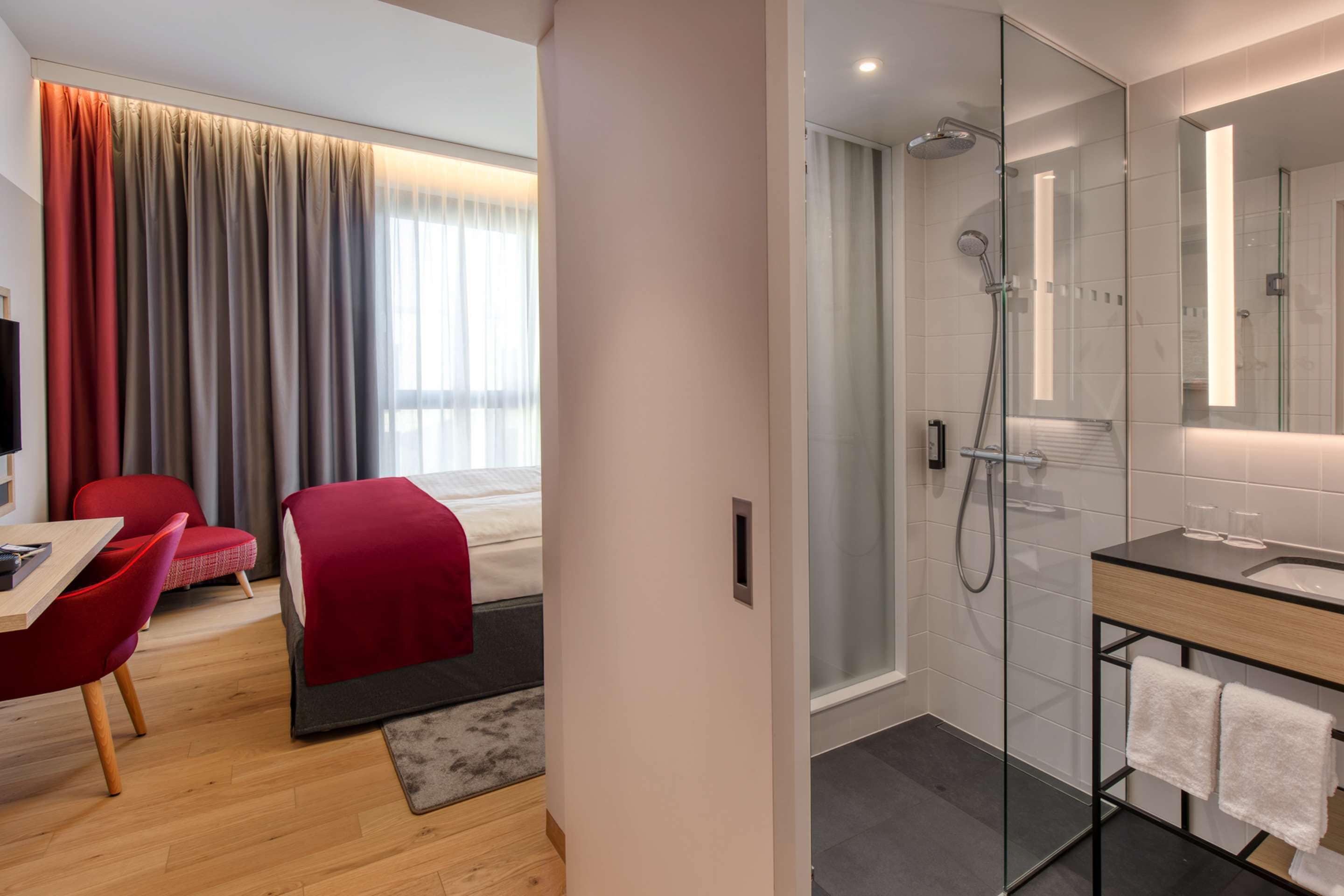 A modern hotel room with a wooden floor, a red chair, a bed with a red blanket, a glass shower enclosure, and a bathroom with a large mirror and a wooden vanity.