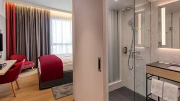 A modern hotel room with a wooden floor, a red chair, a bed with a red blanket, a glass shower enclosure, and a bathroom with a large mirror and a wooden vanity.