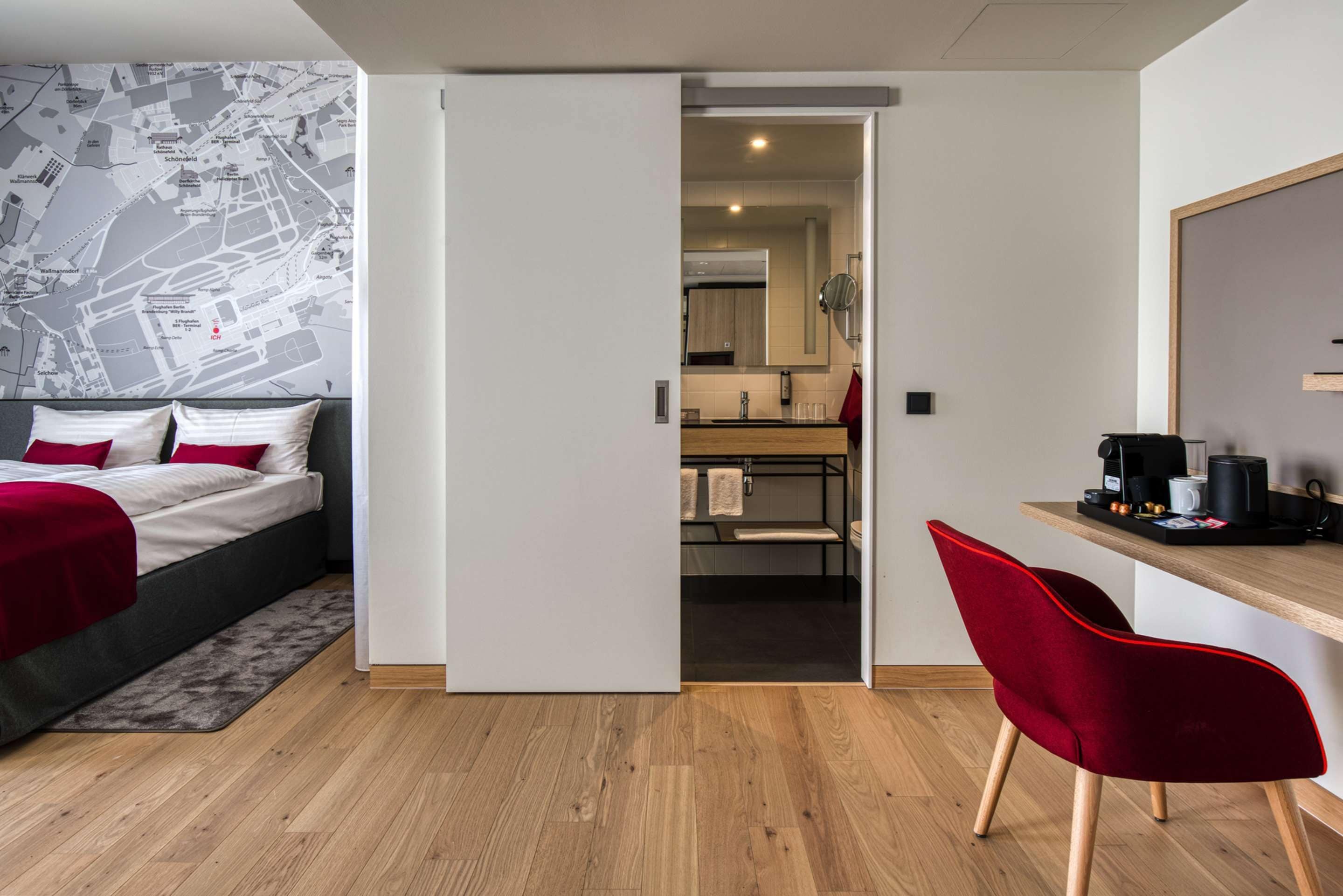 A compact hotel room with a bed, a red chair, a wooden desk, and a kitchenette.