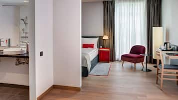 Habitación de hotel con baño, cama con almohadas rojas, una silla roja, un escritorio con lámpara y vistas al exterior a través de amplios ventanales.