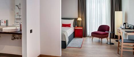 Habitación de hotel con baño, cama con almohadas rojas, una silla roja, un escritorio con lámpara y vistas al exterior a través de amplios ventanales.