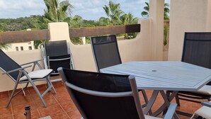 Una terrazza con un tavolo rotondo e quattro sedie, un piccolo barbecue e vista su palme e colline.