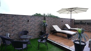 Une terrasse sur le toit avec un mur en briques, une plateforme en bois, une table ronde, deux chaises, un fauteuil, un parasol et des plantes en pot.