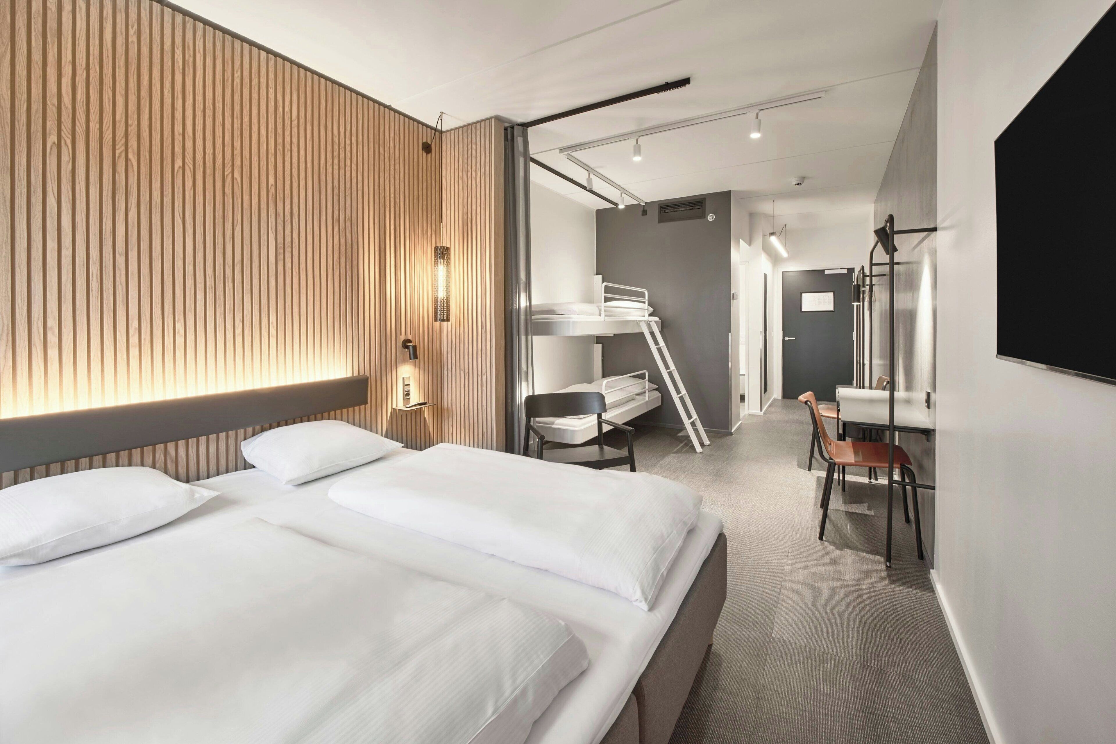 A modern hotel room with a large bed, a wooden accent wall, a desk with a chair, and a bunk bed.