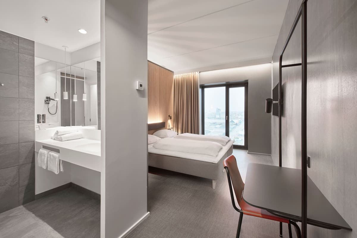 A modern hotel room with a large bed, a separate bathroom with a bathtub, a dining table, and a city view through the window.