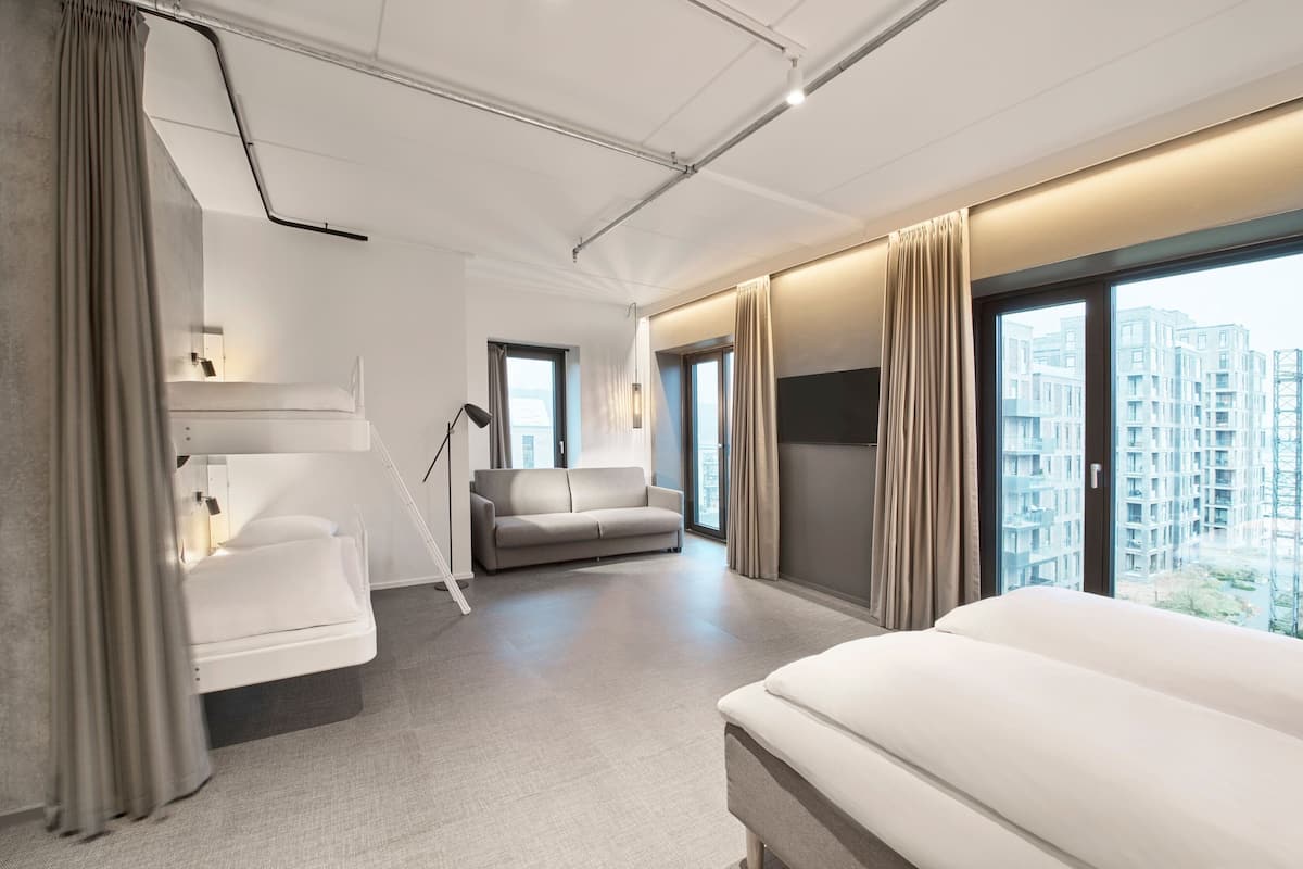 A modern hotel room with a large bed, a sofa, and a desk. There are large windows with curtains, and the room has a city view.
