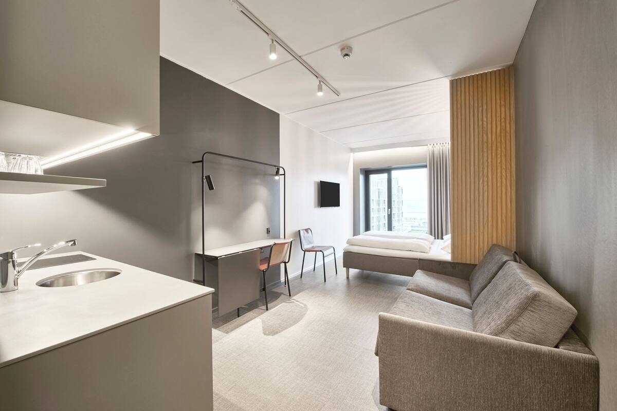 A modern hotel room with a kitchenette, a bed, a desk, and a large window.