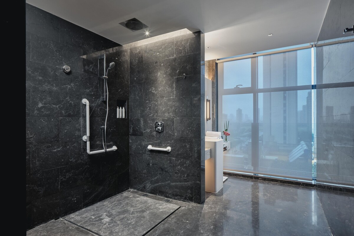A modern bathroom with a shower, grab bars, and a large window offering a city view.