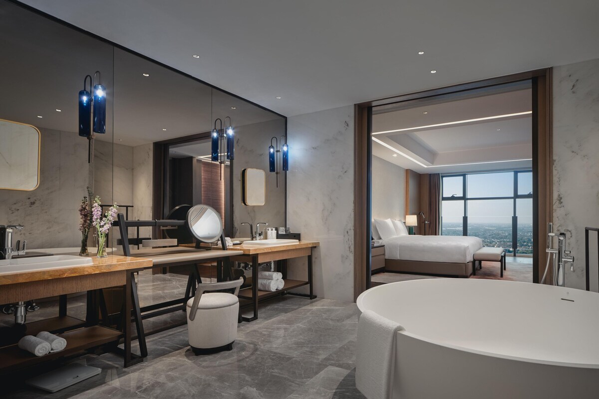 A modern hotel bathroom with a large bathtub, a vanity with a mirror, and a view of the city through large windows.