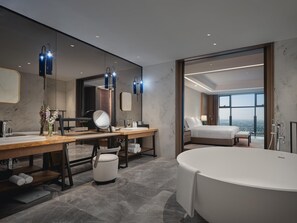 Un baño de hotel moderno con una bañera grande, un tocador con espejo y vistas de la ciudad a través de amplias ventanas.