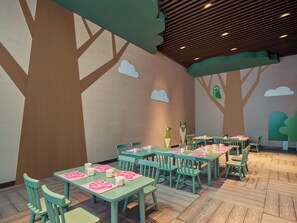 Un salón de juegos para niños con mesas y sillas, suelo de madera y murales de árboles y nubes.