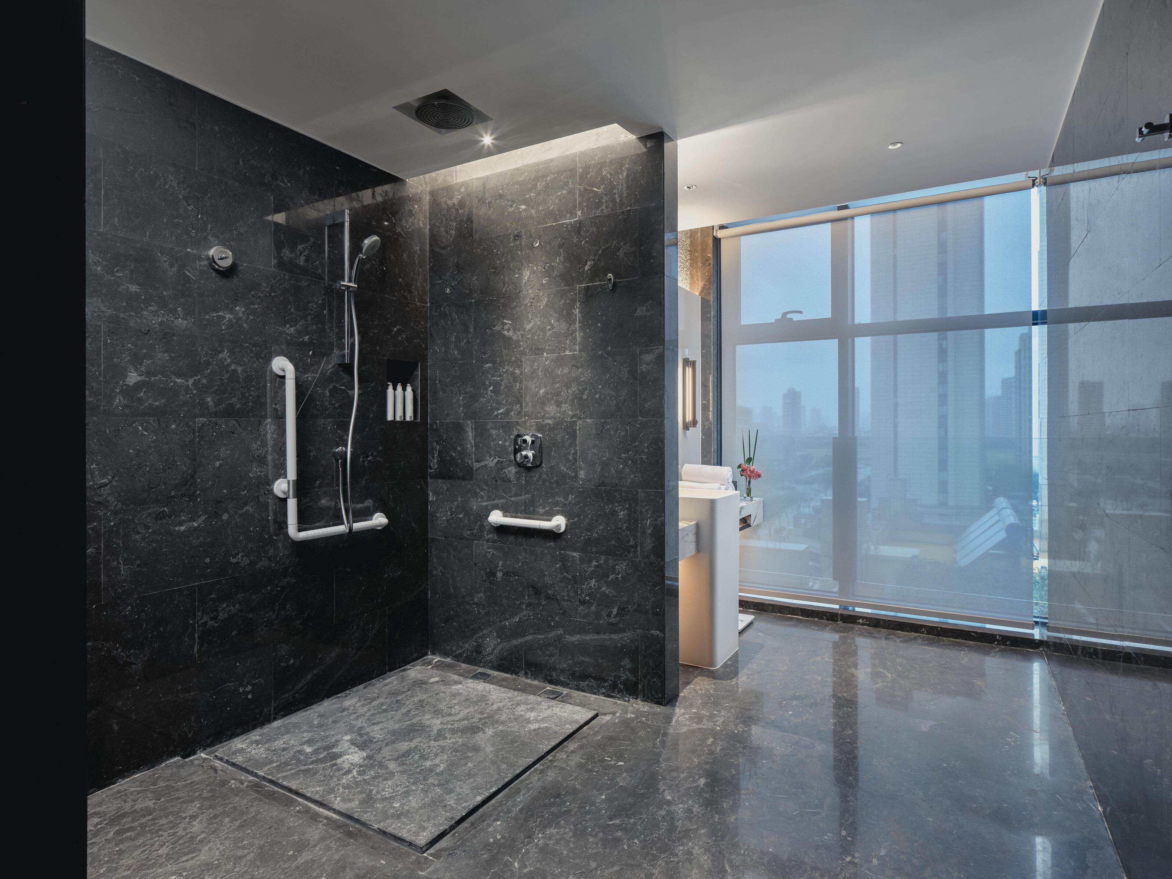 Un baño moderno con ducha, barras de apoyo y un amplio ventanal con vistas a la ciudad.