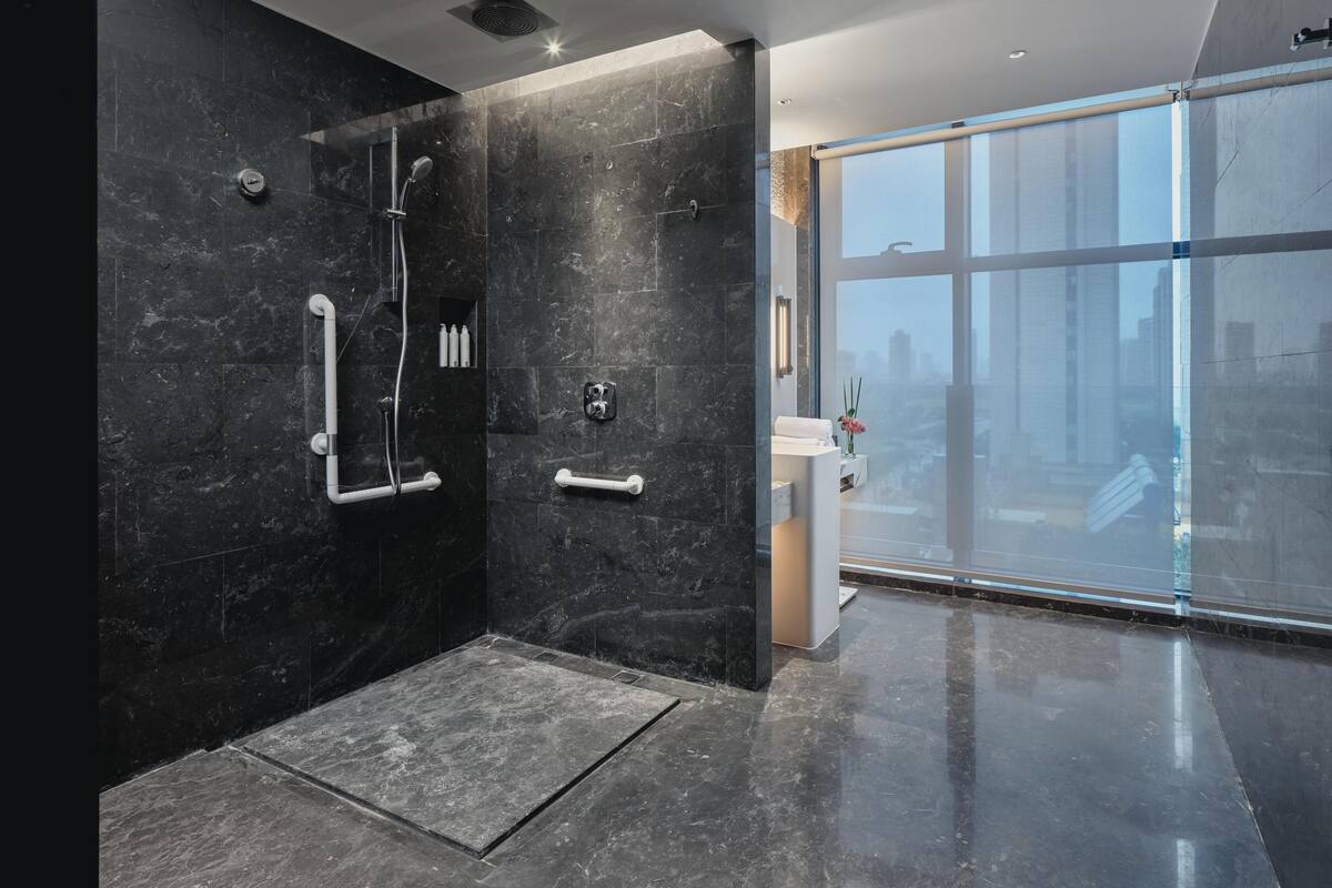 Une salle de bain moderne équipée d’une douche, de barres d’appui et d’une grande fenêtre donnant sur la ville.