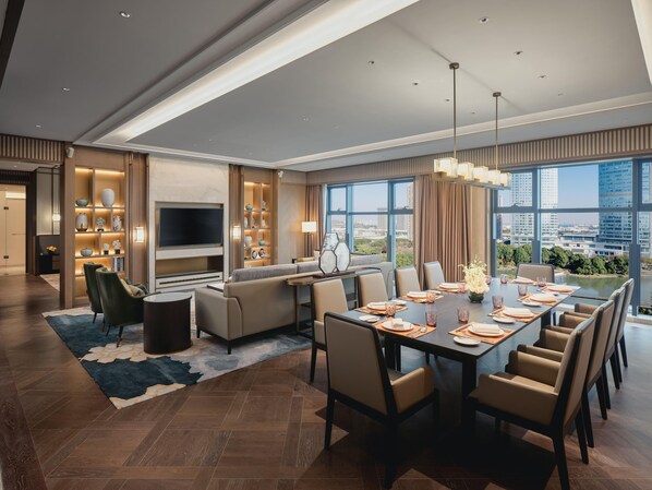 Una habitación de hotel moderna con zona de comedor, sofá, televisor y un amplio ventanal con vista a la ciudad.
