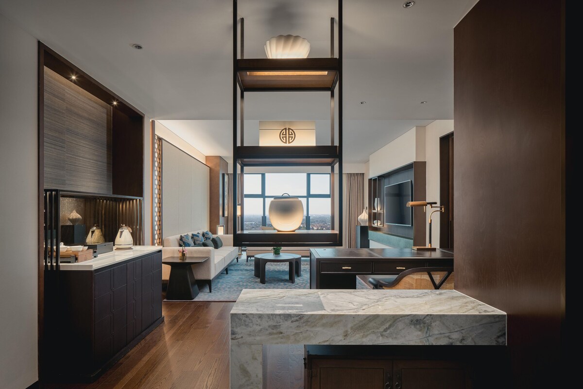 A modern hotel room with a marble kitchen island, a sofa, a dining table, and a large window with a city view.