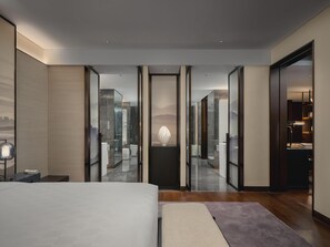 A modern hotel room with a large bed, a desk, and a view through a glass door to a bathroom.