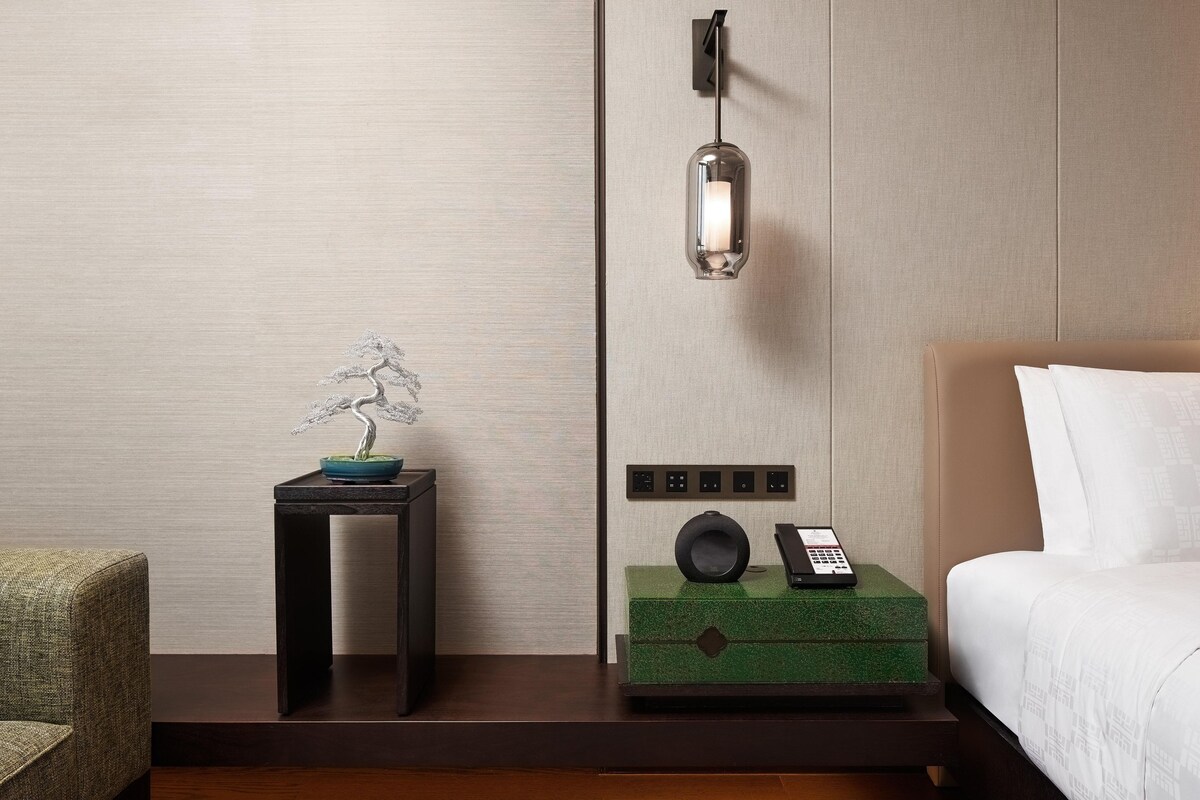 A hotel room with a bed, a bedside table with a phone, a wall-mounted light, and a decorative bonsai tree.