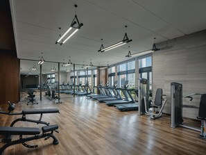 A gym with treadmills, benches, and exercise equipment, large windows, and a wooden floor.