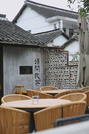 Um edifício tradicional com telhado de azulejos, parede com padrão de tijolos e um letreiro com caracteres chineses. Cadeiras e mesas de vime dispostas no exterior.