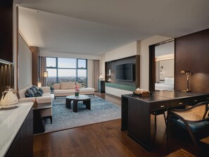 A modern hotel room with a large window, a flat-screen TV, a sofa, a coffee table, a desk, and a chair.