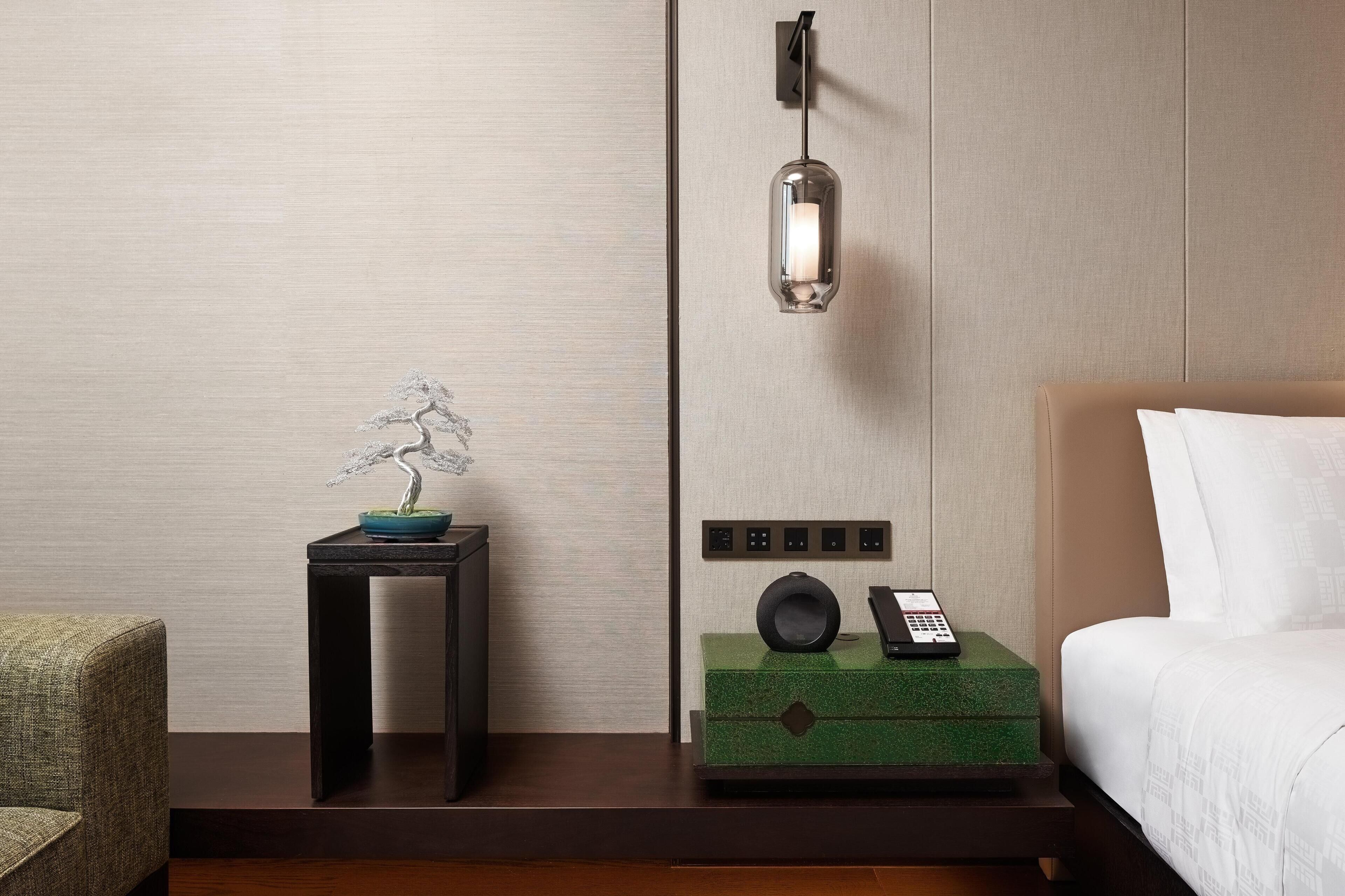 Habitación de hotel con cama, mesita de noche con teléfono, lámpara de pared y un bonsái decorativo.