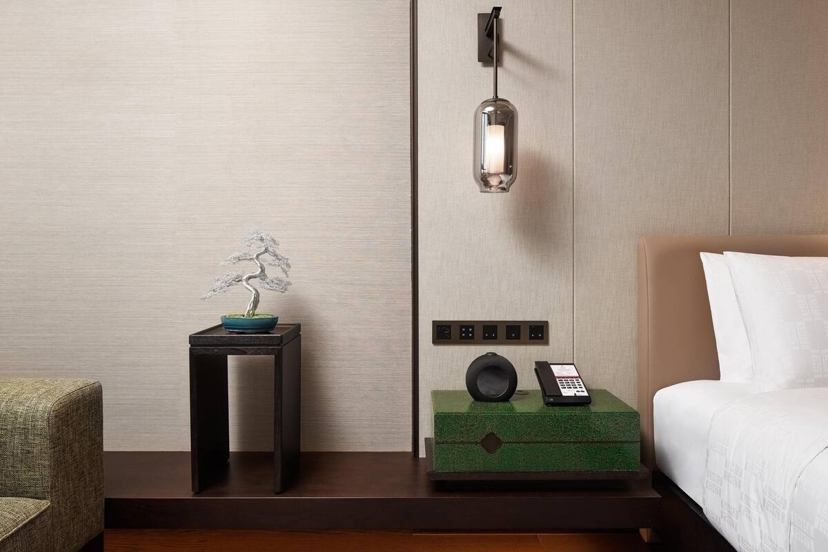 Une chambre d’hôtel avec un lit, une table de chevet avec un téléphone, une lampe fixée au mur et un petit arbre décoratif en bonsaï.