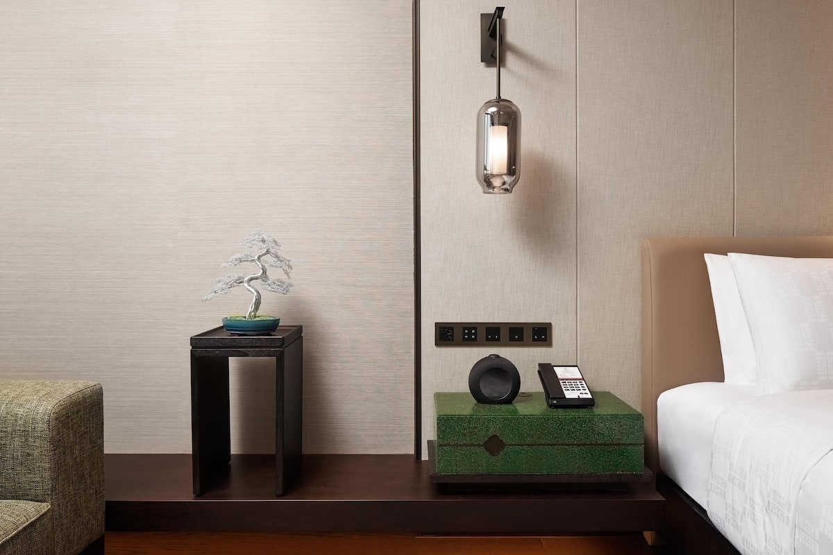 Una camera d'albergo con un letto, un comodino con il telefono, una lampada a parete e un bonsai decorativo.