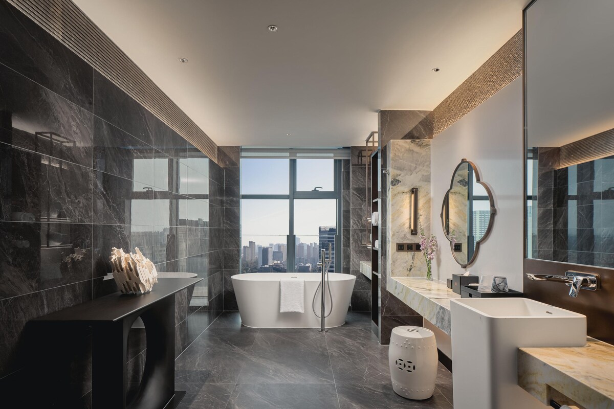 A modern bathroom with a large bathtub, a vanity with a mirror, and a view of the city.